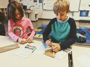 two students work on their landfill solutions environmental engineering challenge. Both students are building their landfills on top of a cardboard base and sculpting clay into cupl ike shapes.